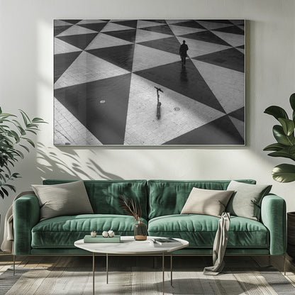 A high-angle, black and white photograph of a man in a coat and fedora walking away across a vast plaza with a geometric pattern of black and light gray triangles. An electric scooter stands alone in the foreground, creating a sense of solitude and contrast. The image is presented within a silver frame. Decor
