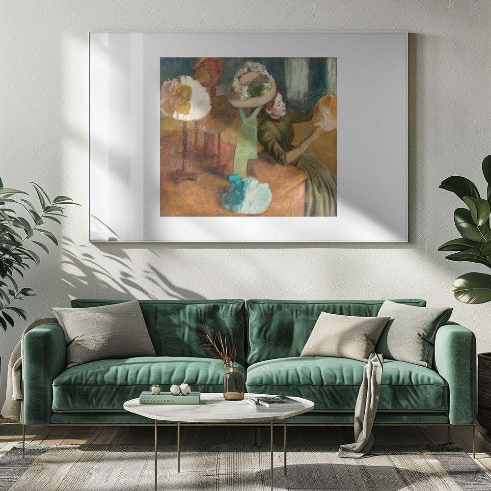 A framed print of Edgar Degas's Impressionist painting 'The Millinery Shop,' showing a woman in a green dress examining a hat in a shop filled with various fashionable hats on display stands. Print