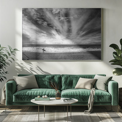 A dramatic black and white photograph of a lone surfer silhouetted against the sea, walking along a sandy beach. The sky is filled with spectacular, radiating clouds that stretch across the horizon, creating a powerful and moody atmosphere. The image is enclosed in a silver frame. Poster