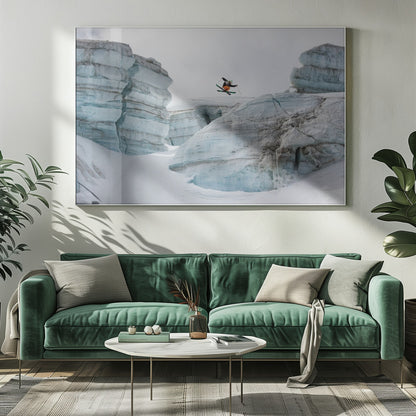 A framed photo of a skier in mid-air, jumping over a snowy crevasse between large, layered glacier formations under an overcast sky. The skier wears a black top and orange pants. Wall Art