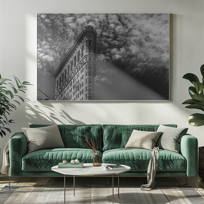 A dramatic low-angle black and white photograph of the Flatiron Building in New York City, with its sharp, triangular corner pointing towards a sky filled with fluffy, white clouds. The image is framed with a brushed silver border. Print