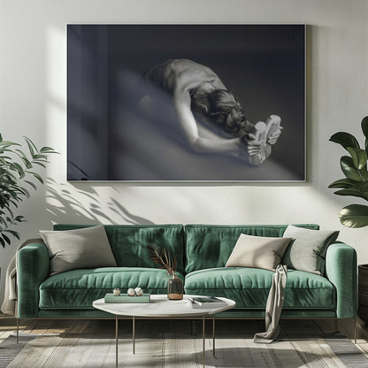 A black and white photograph of a ballerina stretching. She is seated on the floor in a deep forward fold, her torso bent over her legs as she holds onto her pointe shoes. The lighting accentuates the musculature of her back and shoulders, and her hair is styled in a braided bun. Decor