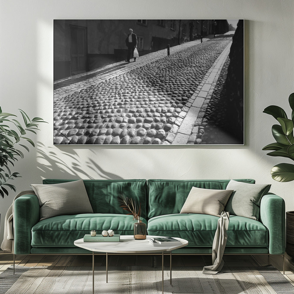 A framed, black and white photograph capturing an elderly man in a suit walking up a steep cobblestone street. The man carries a white bag and casts a long shadow behind him in the harsh sunlight. The street is lined with old, multi-story buildings, creating a classic European city scene. Poster