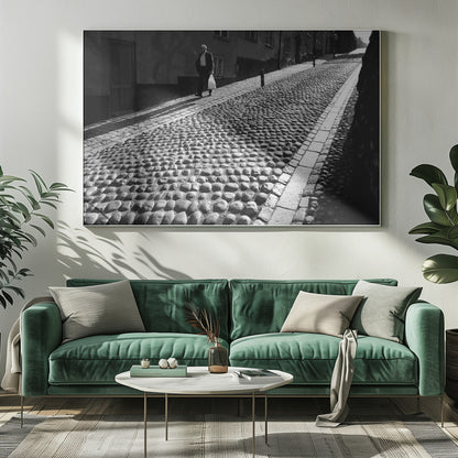 A framed, black and white photograph capturing an elderly man in a suit walking up a steep cobblestone street. The man carries a white bag and casts a long shadow behind him in the harsh sunlight. The street is lined with old, multi-story buildings, creating a classic European city scene. Poster