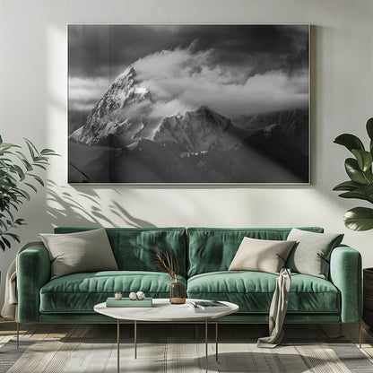 A dramatic black and white photograph of a snow-covered mountain peak being enveloped by thick, swirling clouds under a dark, stormy sky. The rugged slopes and sharp ridges of the mountain are highlighted by the snow, while a dark forest sits at the base. The entire image is presented within a textured silver frame. Print