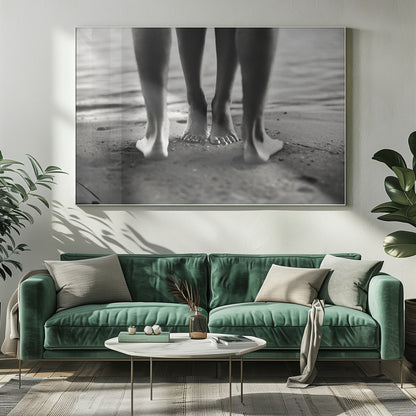 A framed, black and white photograph showing a low-angle view of two people's bare feet and lower legs standing on a wet sandy beach. The central pair of feet is in focus, while another person's legs in the foreground are blurred, creating depth. The water's edge is visible in the background. Artwork
