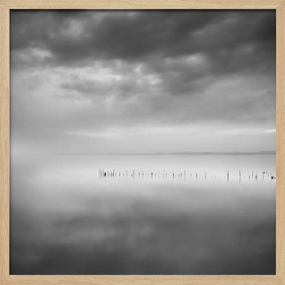 A minimalist black and white photograph in a light wood frame, depicting a calm, serene lake under a heavy, cloudy sky. The still water reflects the sky, with a row of weathered wooden posts breaking the surface along the horizon. Print