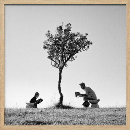 A framed, black and white photograph depicting a surreal scene of a man and a child sitting on toilets in an open grassy field under a single tree, both wearing hats and reading. Print