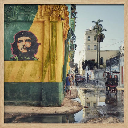 A framed photograph of a vibrant street corner in a city like Havana, Cuba, with a large mural of Che Guevara on a yellow and green wall. The wet street reflects the buildings and a palm tree, while people, a vintage car, and a rickshaw occupy the narrow road. Poster