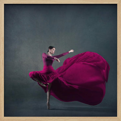 A graceful ballet dancer in a vibrant magenta dress strikes a pose on pointe, with the long, flowing fabric of her skirt billowing out dramatically behind her against a dark, textured gray background, all enclosed in a simple light wood frame. Decor