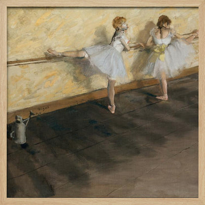 A framed impressionist painting by Edgar Degas depicting two ballet dancers in white tutus practicing at a barre. The studio has a yellow wall and a dark brown wooden floor. One dancer stretches her leg on the barre while the other stands beside her. Artwork