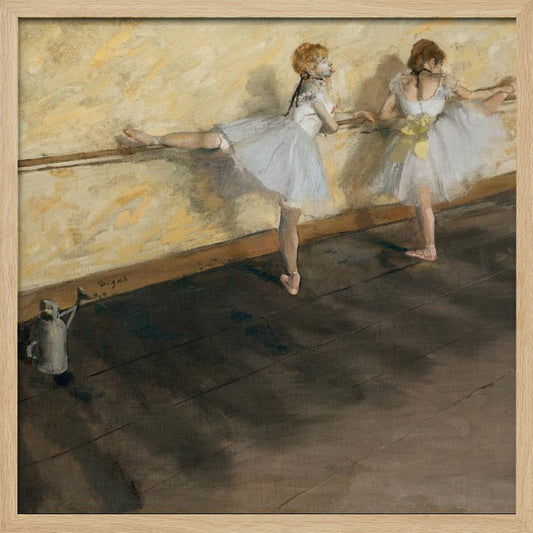 A framed impressionist painting by Edgar Degas depicting two ballet dancers in white tutus practicing at a barre. The studio has a yellow wall and a dark brown wooden floor. One dancer stretches her leg on the barre while the other stands beside her. Artwork