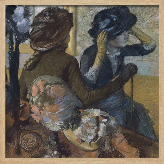 A framed impressionist painting by Edgar Degas, 'The Millinery Shop', depicting two women. One woman, seen from the back, wears a brown coat and hat, while the other tries on a dark blue hat, adjusting it with her gloved hands. Colorful hats are displayed in the foreground. Print