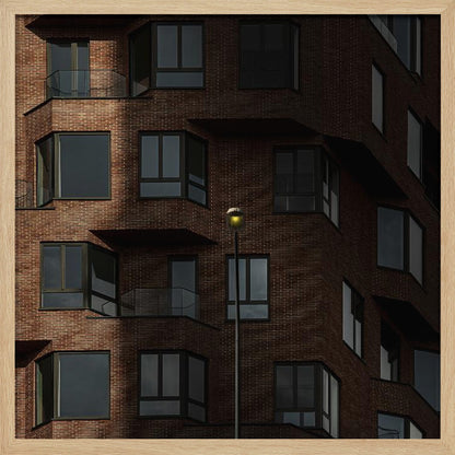 A close-up photograph of a modern red-brick apartment building, framed in light wood. A sharp shadow line divides the facade, with a lit streetlamp providing a warm yellow glow in the shaded area. Decor