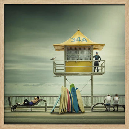 A vintage-style photograph of a yellow lifeguard tower labeled '34A' on a beach boardwalk under an overcast, greenish sky. A lifeguard in a wetsuit stands on the balcony, while a collection of colorful surfboards leans against the base. People are seen relaxing on benches nearby. Print