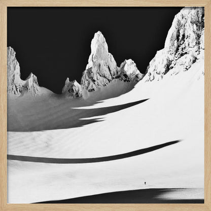 A framed, high-contrast black and white photograph of a majestic, snow-covered mountain range. Jagged peaks are sunlit against a stark black sky, casting long, dramatic shadows across a vast, pristine snowfield below. A tiny, solitary figure stands in the snow, highlighting the immense scale of the landscape. Print