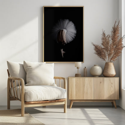 A dramatic photograph of a ballerina against a solid black background. She is bent completely forward, showing her arched back to the camera. Her white tutu fans out in a wide circle around her, and one foot in a pointe shoe is visible beneath her, balanced on its tip. Print