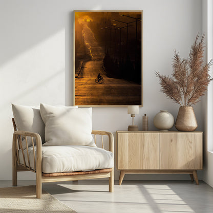 A silhouette of a person riding a bicycle across a wide, winding road illuminated by the warm, golden light of a sunrise or sunset. The road stretches uphill into the distance, and tall streetlights stand in shadow on the right. Artwork