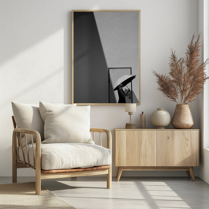A minimalist black and white photograph of a woman in a black dress holding a two-toned umbrella. She stands in front of a door against a light wall, with a sharp, diagonal shadow dramatically cutting across the upper left of the image, creating a high-contrast, geometric composition. Decor