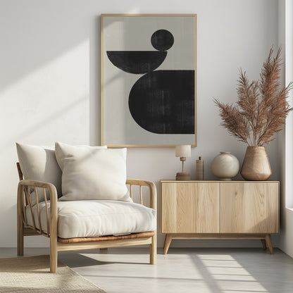 A minimalist abstract artwork featuring three charcoal black shapes stacked against a light beige textured background. At the bottom is a large, rounded shape, on which a semi-circle is balanced. A smaller, full circle rests on top of the semi-circle, creating a sense of precarious balance. The piece is enclosed in a simple black frame. Decor