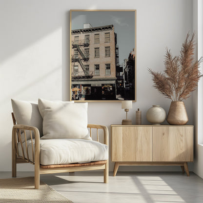 A low-angle, desaturated photograph of a classic brown brick building in a city, featuring multiple windows and a black metal fire escape zigzagging up the side. The sky is a pale, hazy blue, and the ground floor has dark storefronts. Decor