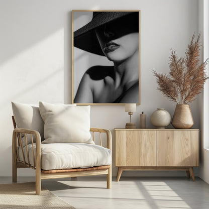 A dramatic black and white close-up photograph of a woman wearing a large, dark, wide-brimmed hat. The hat casts a deep shadow over the upper half of her face, obscuring her eyes and creating a sense of mystery. Only her nose, full lips, chin, neck, and shoulder are visible, highlighted by a strong light source. Decor