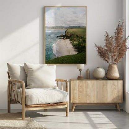 A high-angle photograph of a secluded tropical beach, with turquoise waves gently washing onto the white sand. A lush green cliff covered in grass and small trees frames the cove, with distant rolling hills visible under a soft, cloudy sky. Decor