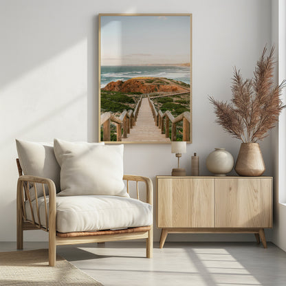 A wooden staircase and walkway descends a green, scrub-covered hill towards a rocky point overlooking a churning blue ocean under a pale, hazy sky. Print