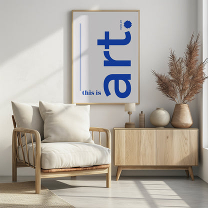 A minimalist art print in a black frame, featuring a white background with blue text. The word 'art' is written vertically in a large, bold lowercase font. To the left, smaller text reads 'this is'. At the top right, 'hello art' is written next to a blue starburst graphic. Wall Art