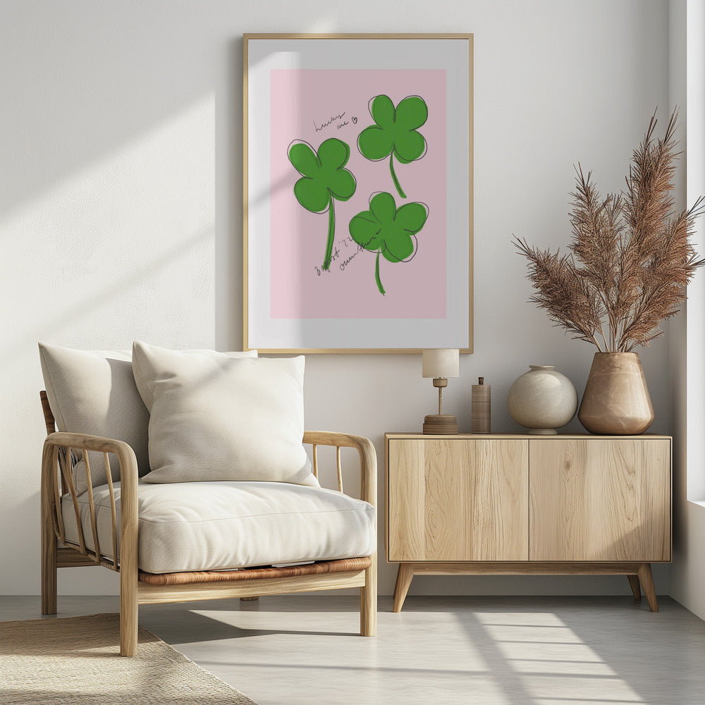 A framed art print featuring three hand-drawn four-leaf clovers in green with a loose black outline, set against a solid light pink background. Cursive handwriting on the print reads 'lucky me ♡' and includes a signature. Artwork