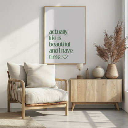 A framed poster with a white background and a black frame. The poster features text in a green, lowercase, sans-serif font that reads, 'actually, life is beautiful and i have time.' followed by an outlined heart symbol. Decor
