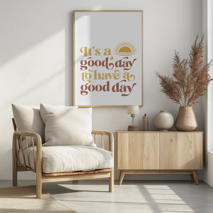 A framed art print with a white background featuring the quote 'It's a good day to have a good day' in a retro, curvy font. The text color alternates between terracotta brown and mustard yellow. Above the text is a small yellow icon of a sun rising over waves. Wall Art
