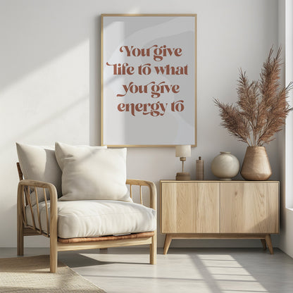 A framed motivational poster featuring the quote 'You give life to what you give energy to'. The text is printed in a dusty rose color using a stylish, retro serif font on a light gray background with subtle wavy shapes. Poster