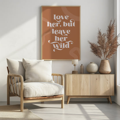 A framed typography poster with a terracotta brown background. In the center, the quote 'love her, but leave her wild' is written in a stylized, decorative, cream-colored font. Faint, lighter brown abstract shapes are visible in the background. Decor