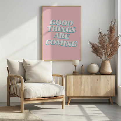 A framed poster with a light pink background featuring the motivational phrase 'GOOD THINGS ARE COMING'. The text is in a white, retro-style serif font with a light blue drop shadow, giving it a three-dimensional effect. Poster