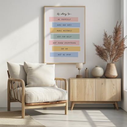 A framed inspirational poster with a white background. At the top, in a black handwritten font, it says 'it's okay to...'. Below is a list of affirmations, each on a different colored pastel rectangle: 'BE YOURSELF' on pink, 'HAVE BAD DAYS' on blue, 'MAKE MISTAKES' on yellow, 'ASK FOR HELP' on green, 'NOT KNOW EVERYTHING' on pink, 'SAY NO' on yellow, and 'NOT BE OKAY' on blue. Poster