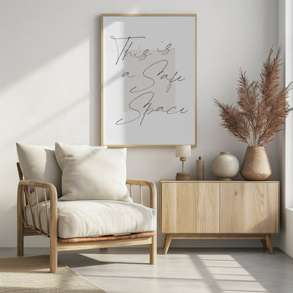 A minimalist art print in a black frame with the words 'This is a Safe Space' written in a flowing black cursive script over a pale beige rectangle on a white background. Artwork