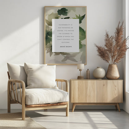 A framed art print featuring an inspirational quote by Brené Brown. The quote, 'Vulnerability is not winning or losing. It's having the courage to show up when you can't control the outcome,' is printed in black uppercase letters on a central white rectangle. The background consists of a soft watercolor illustration of green and beige leaves. The piece is enclosed in a simple black frame. Wall Art