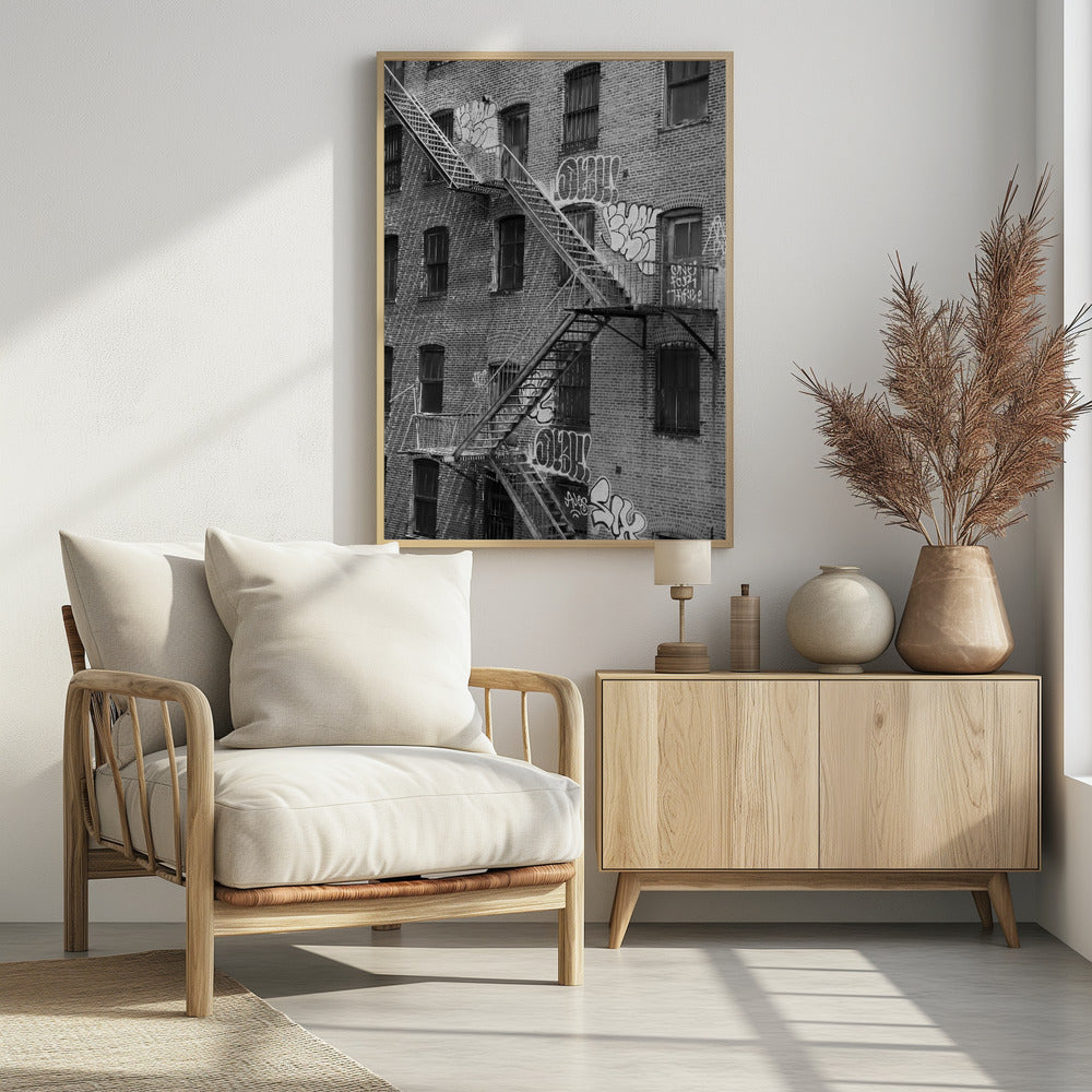 A black and white photograph of a brick apartment building with a prominent metal fire escape zigzagging across its facade. The walls are adorned with various styles of graffiti, and the windows are covered with bars, creating a gritty, urban scene. Decor