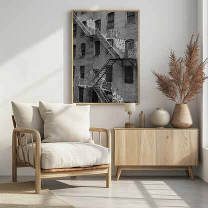 A black and white photograph of a brick apartment building with a prominent metal fire escape zigzagging across its facade. The walls are adorned with various styles of graffiti, and the windows are covered with bars, creating a gritty, urban scene. Decor