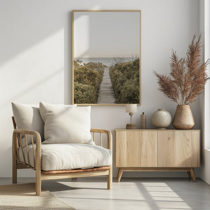 A vertical photograph of a weathered wooden boardwalk leading through dense coastal greenery towards the ocean under a pale, hazy sky, framed in black. Wall Art