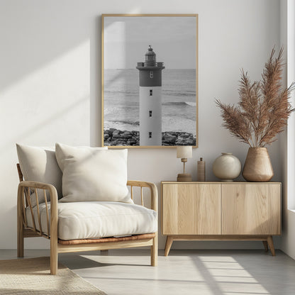 A framed, black and white photograph of a tall, cylindrical lighthouse standing on a rocky shore. The lighthouse is dark grey on its upper section and white on the lower, with the ocean and a grey sky in the background. Print