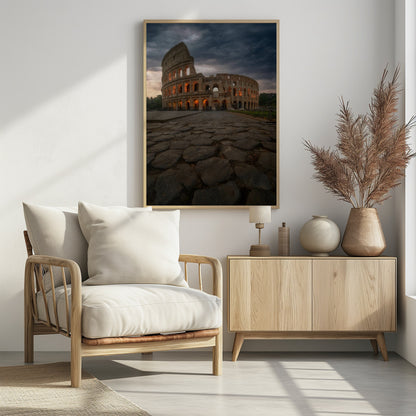 A low-angle view of the Roman Colosseum at dusk, with its arched windows glowing with warm orange light under a dramatic, stormy sky. An ancient cobblestone road leads towards the iconic amphitheater. Print