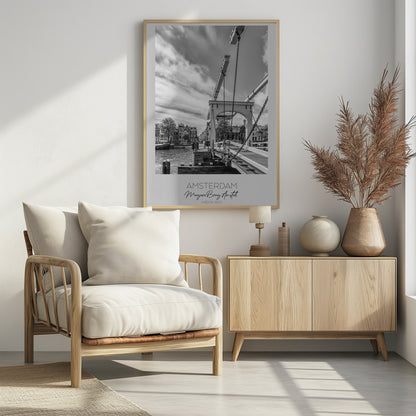 A black and white travel poster of Amsterdam, featuring a side view of the Magere Brug, a traditional wooden drawbridge, over the Amstel river. Classic canal houses line the waterway in the background under a dramatic, cloudy sky. The image is in a black frame and has text at the bottom that reads 'AMSTERDAM', 'Magere Brug, Amstel', and 'NEDERLAND'. Artwork