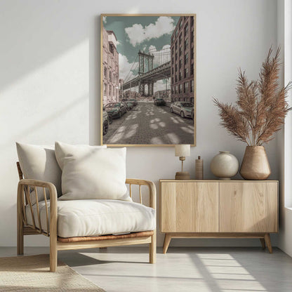 A vertical photograph of the Manhattan Bridge from a cobblestone street in DUMBO, Brooklyn, framed by tall brick buildings. The image has a vintage aesthetic with a muted teal sky and sepia-toned buildings and streets. Print