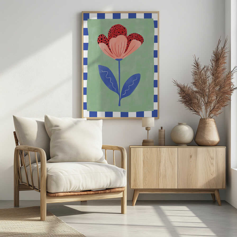 A framed illustration of a stylized flower with red polka-dotted and pink petals. The flower has a blue stem and two blue leaves, set against a sage green background with a blue and white checkered border. Artwork