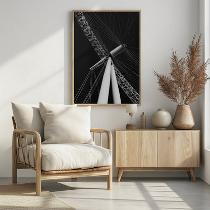 A dramatic low-angle, black and white photograph of a large Ferris wheel, focusing on the central hub and supporting structure. The image captures the white A-frame legs and the intricate lattice of the wheel's rim, with numerous tension cables radiating outwards against a solid black sky, creating a strong sense of perspective and geometric pattern. Artwork