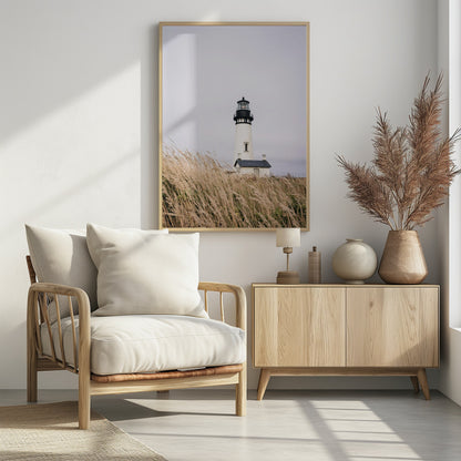 A framed photograph of a white lighthouse with a black top, set against a muted, overcast sky. The foreground is filled with tall, wild beige grass blowing in the wind, partially obscuring the base of the lighthouse. Decor