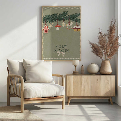 A vintage-style holiday poster with a sage green background. A pine branch hangs at the top, decorated with illustrated ornaments including houses, stars, a mushroom, and a rocking horse. Below, the text 'COZY SEASON' is written in a dark serif font with a light-colored bow underneath. Artwork