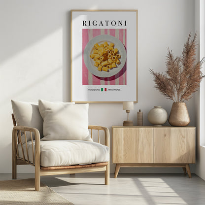 A framed art print of a vintage-style Italian food poster. It features a painting of a white plate full of uncooked rigatoni pasta on a pink and white striped background. The word 'RIGATONI' is printed in large black letters at the top, and 'TRADIZIONE ARTIGIANALE' with a small Italian flag is printed at the bottom. Artwork